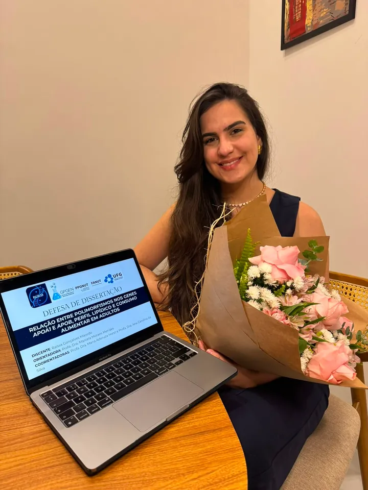 Raissa sorrindo com um buquê de flores ao lado do notebook com a apresentação do mestrado