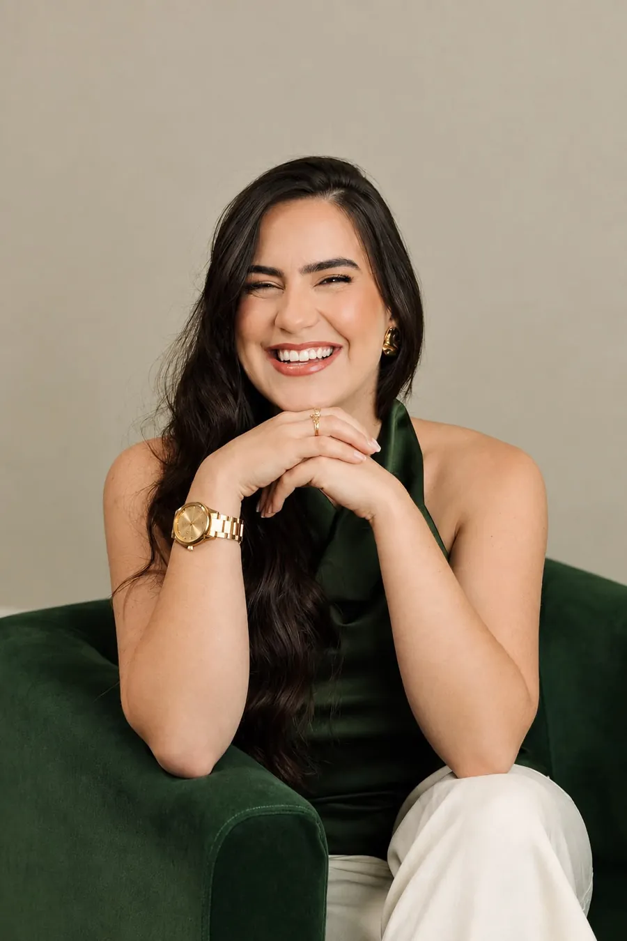 Raissa Macedo rindo, com as mãos apoiadas no queixo, de blusa verde em uma poltrona de veludo verde